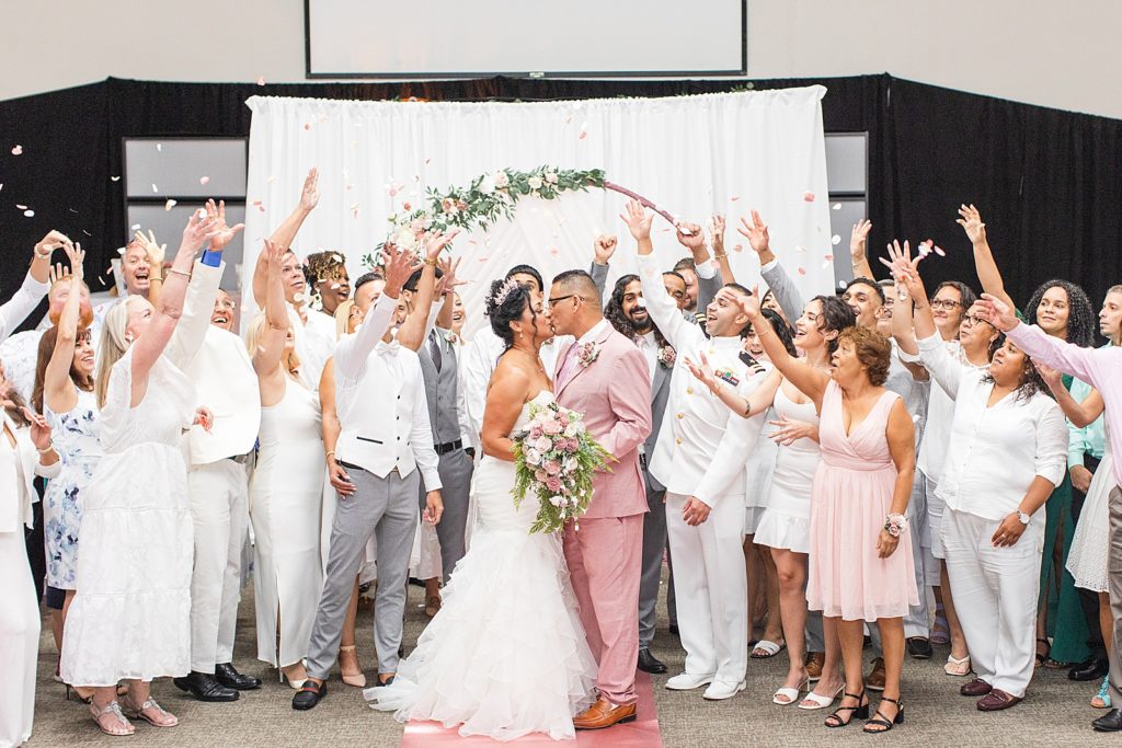 A group picture with the bride and groom kissing while their family throws pink rose petals behind them.
