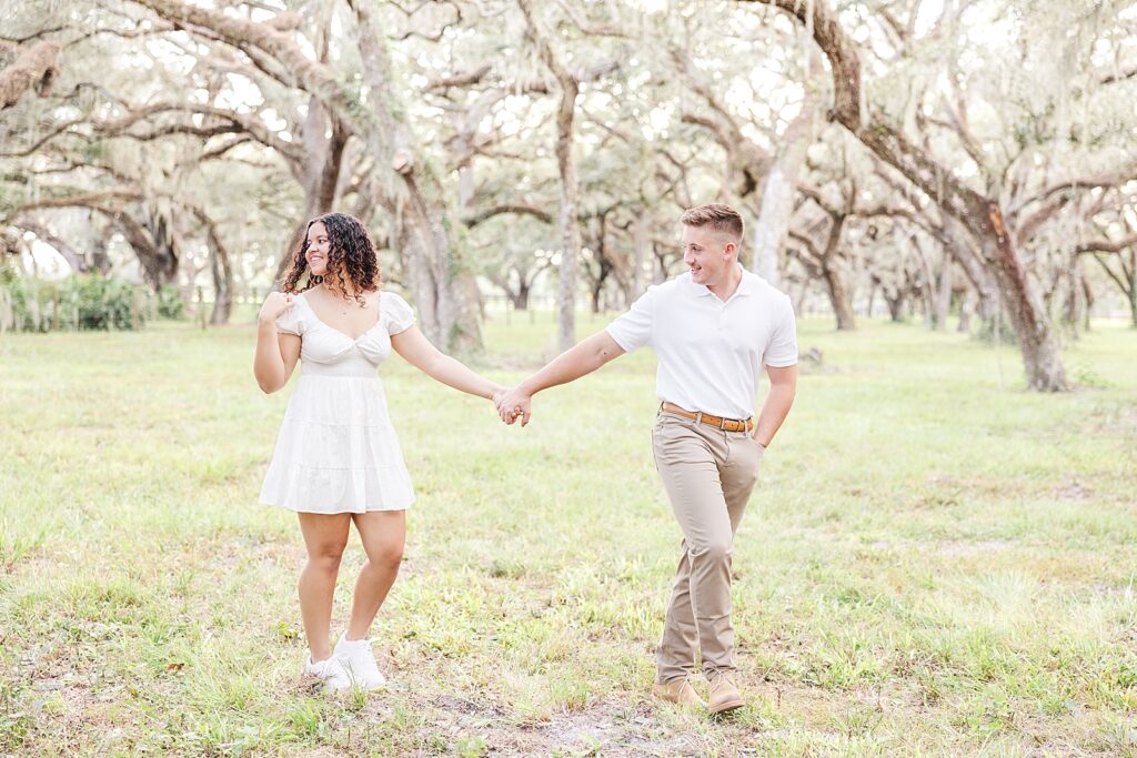A engaged couple walking through a field with mossy oak trees.